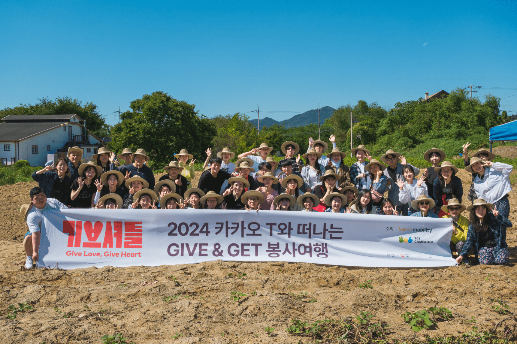 기브셔틀 5회차 참여자들이 '이공이사 카카오 T와 떠나는 기브앤겟 봉사여행' 글자가 적혀 있는 현수막을 든 채 농촌에 모여 단체사진을 촬영하고 있습니다.